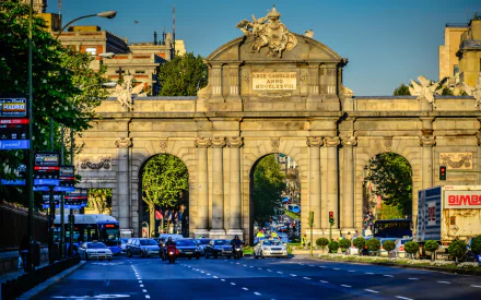HD desktop wallpaper of the man-made Puerta de Alcalá: Neoclassical stone triumphal arch over a busy Madrid avenue at golden hour, with cars and surrounding city buildings.