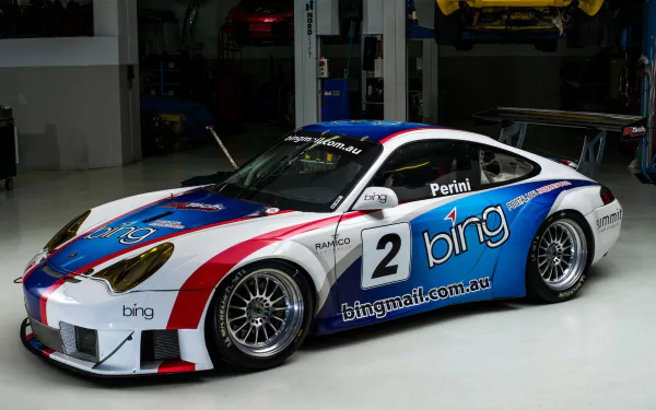 HD desktop wallpaper of a Porsche 911 race car featuring a blue, white, and red livery with sponsor logos, displayed in a garage setting.