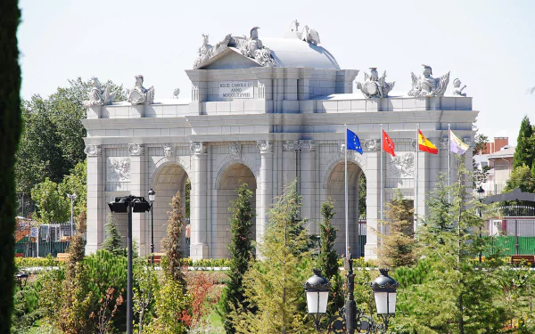 HD PC desktop wallpaper featuring the man-made Puerta de Alcalá monument with flags and greenery in the foreground under a clear sky.