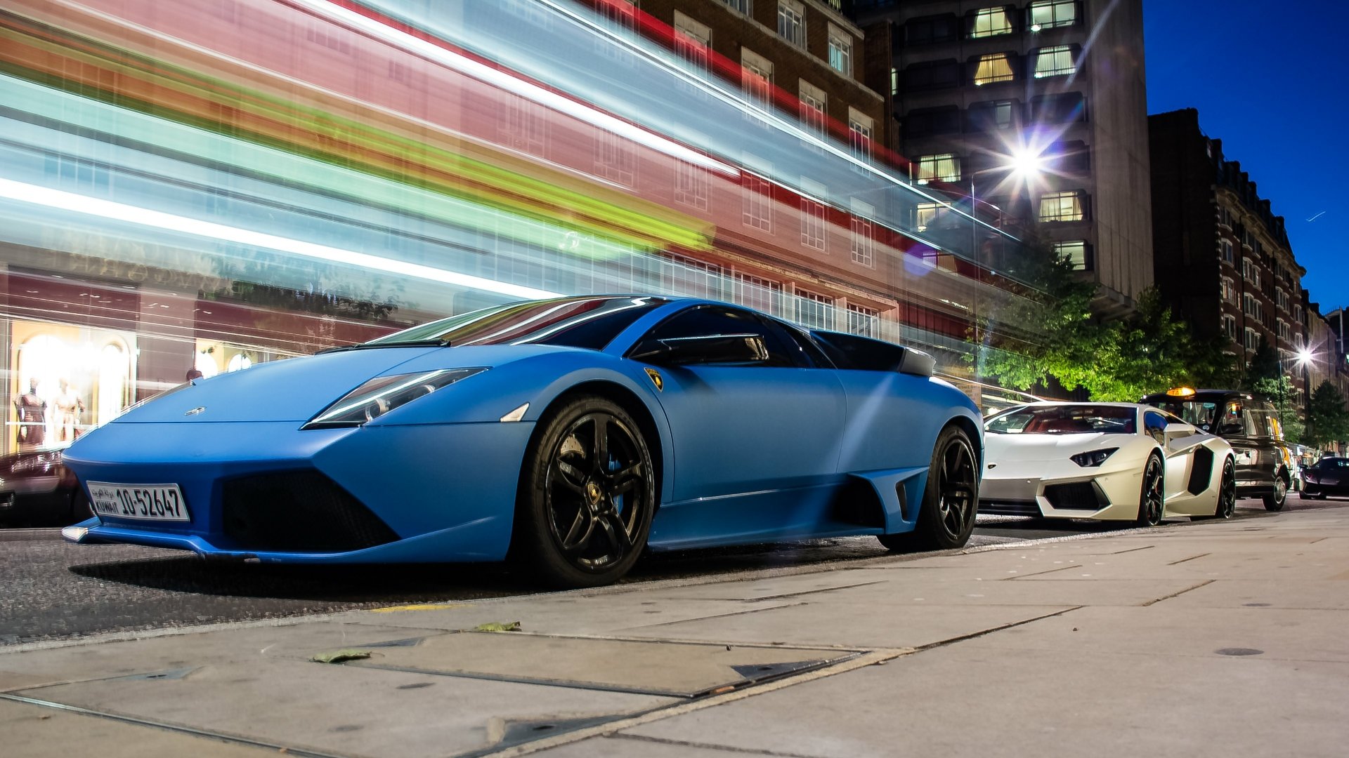 A vibrant 4K Ultra HD wallpaper featuring a blue Lamborghini Murcielago and a white Lamborghini parked on a city street at night.