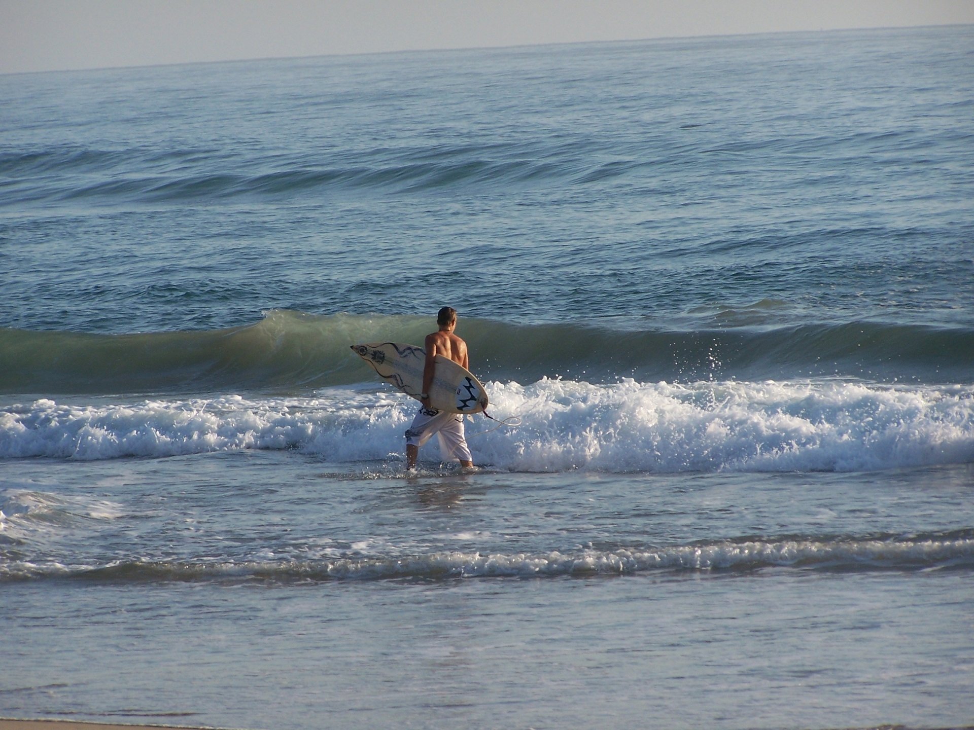 Surfer carrying a surfboard walks into ocean waves, captured in an HD desktop wallpaper showcasing surfing and ocean sports action.
