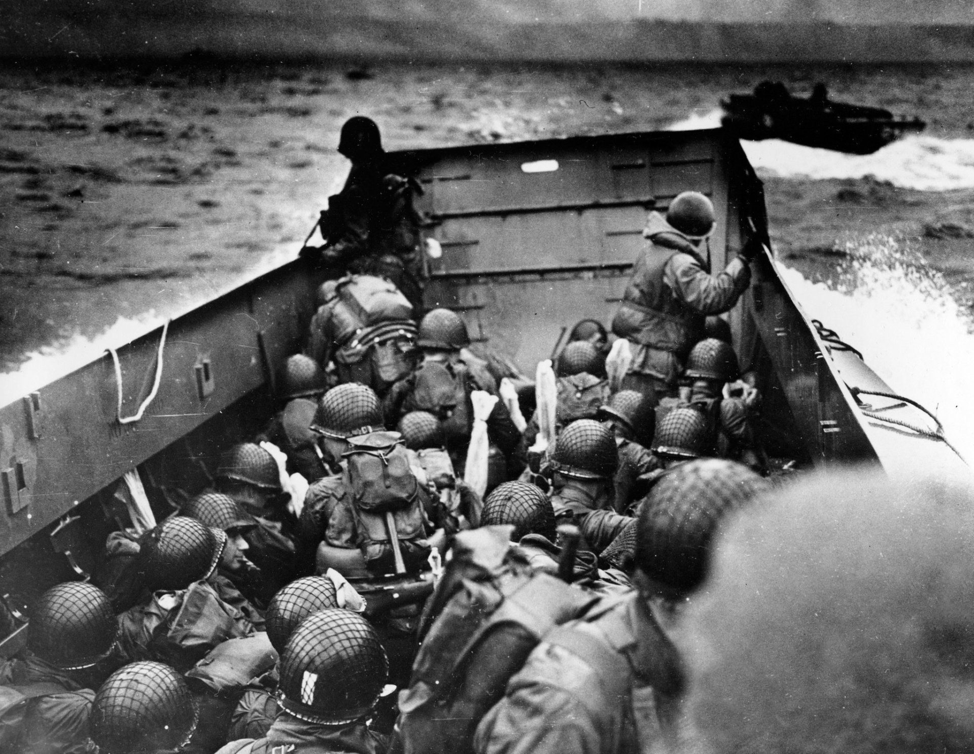 Black and white HD desktop wallpaper depicting armed soldiers in a landing craft approaching the shore during the D-Day military invasion.