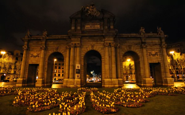 HD PC desktop wallpaper and background: illuminated Puerta de Alcalá at night — a man-made stone archway framed by flowerbeds and warm streetlights.