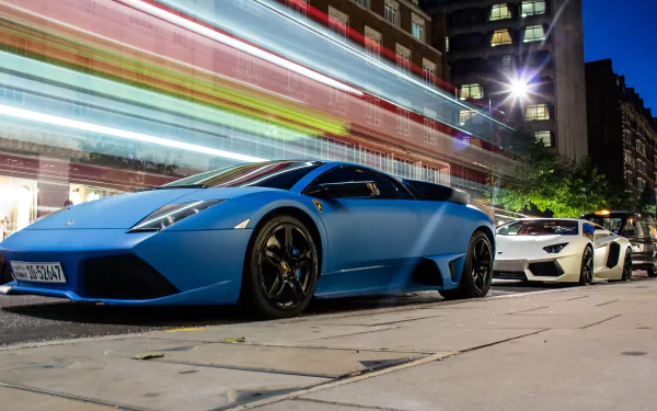 A vibrant 4K Ultra HD wallpaper featuring a blue Lamborghini Murcielago and a white Lamborghini parked on a city street at night.
