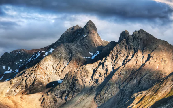 Majestic alpine peaks rise dramatically under a cloudy sky, showcasing the stunning beauty of nature in the Alps. This HD wallpaper captures their rugged terrain and vibrant light.