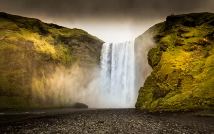 HD desktop wallpaper showcasing a powerful waterfall cascading between moss-covered cliffs under a muted sky, highlighting nature's raw beauty.