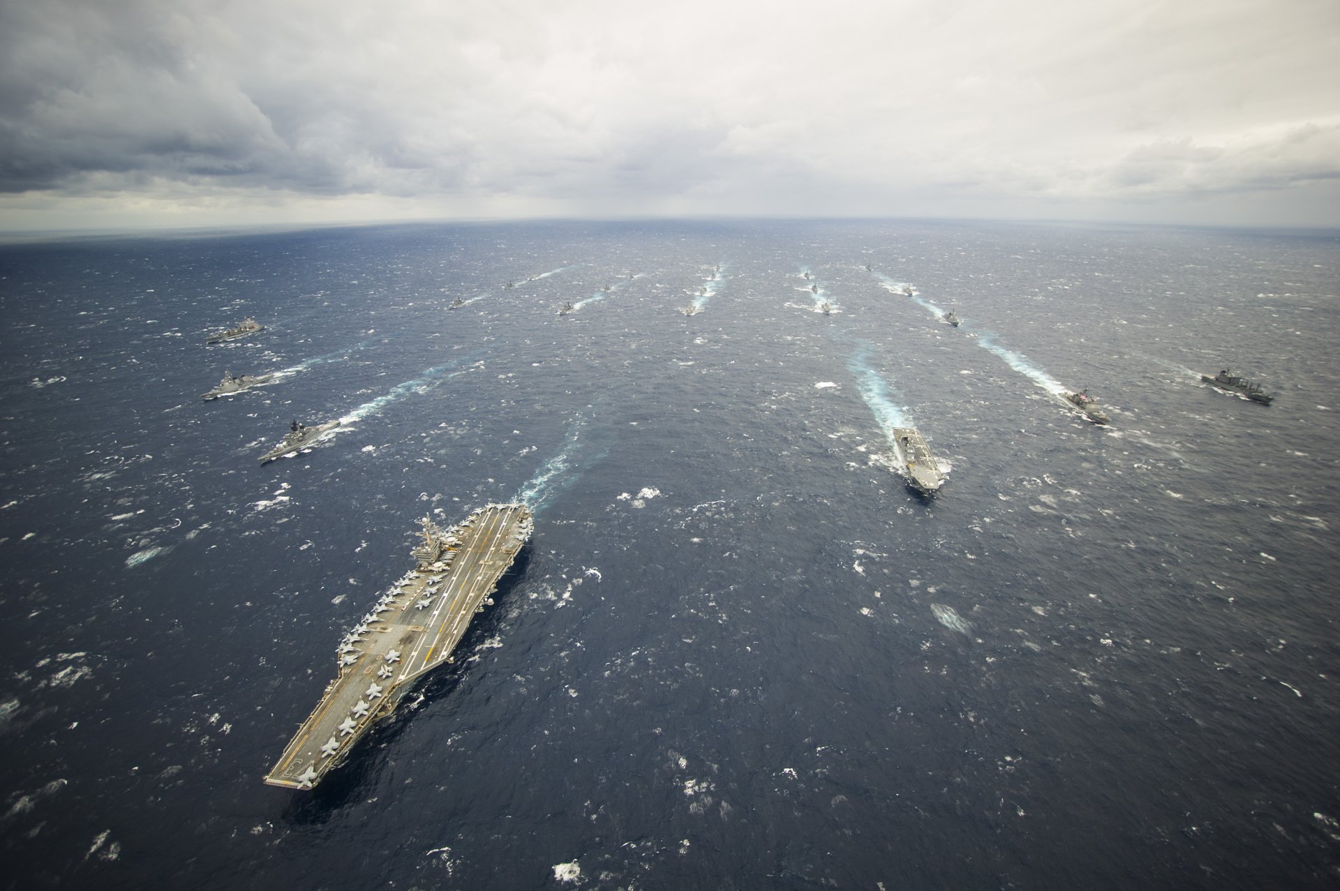 A fleet of navy warships, led by the aircraft carrier USS George Washington (CVN-73), sails across the ocean under a cloudy sky.