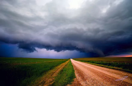 HD PC desktop wallpaper: a gravel road through green fields leading into a dramatic storm, dark rain clouds filling the sky in a moody nature scene.