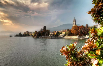Scenic view of blossoming plants along the shoreline in Pallanza, Verbania, Italy, with historic buildings and a church tower under a dramatic sky.