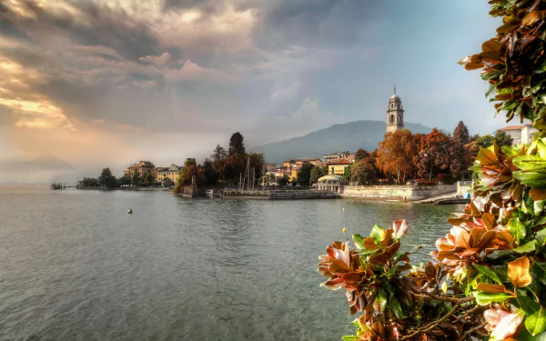 Scenic view of blossoming plants along the shoreline in Pallanza, Verbania, Italy, with historic buildings and a church tower under a dramatic sky.