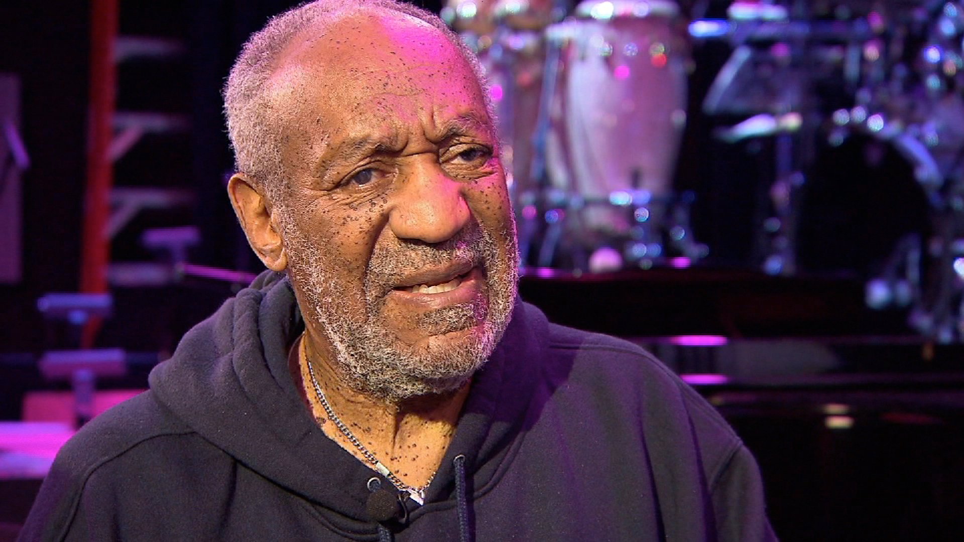 A HD desktop wallpaper featuring a close-up of a man wearing a hoodie with a musical drum set in the background.