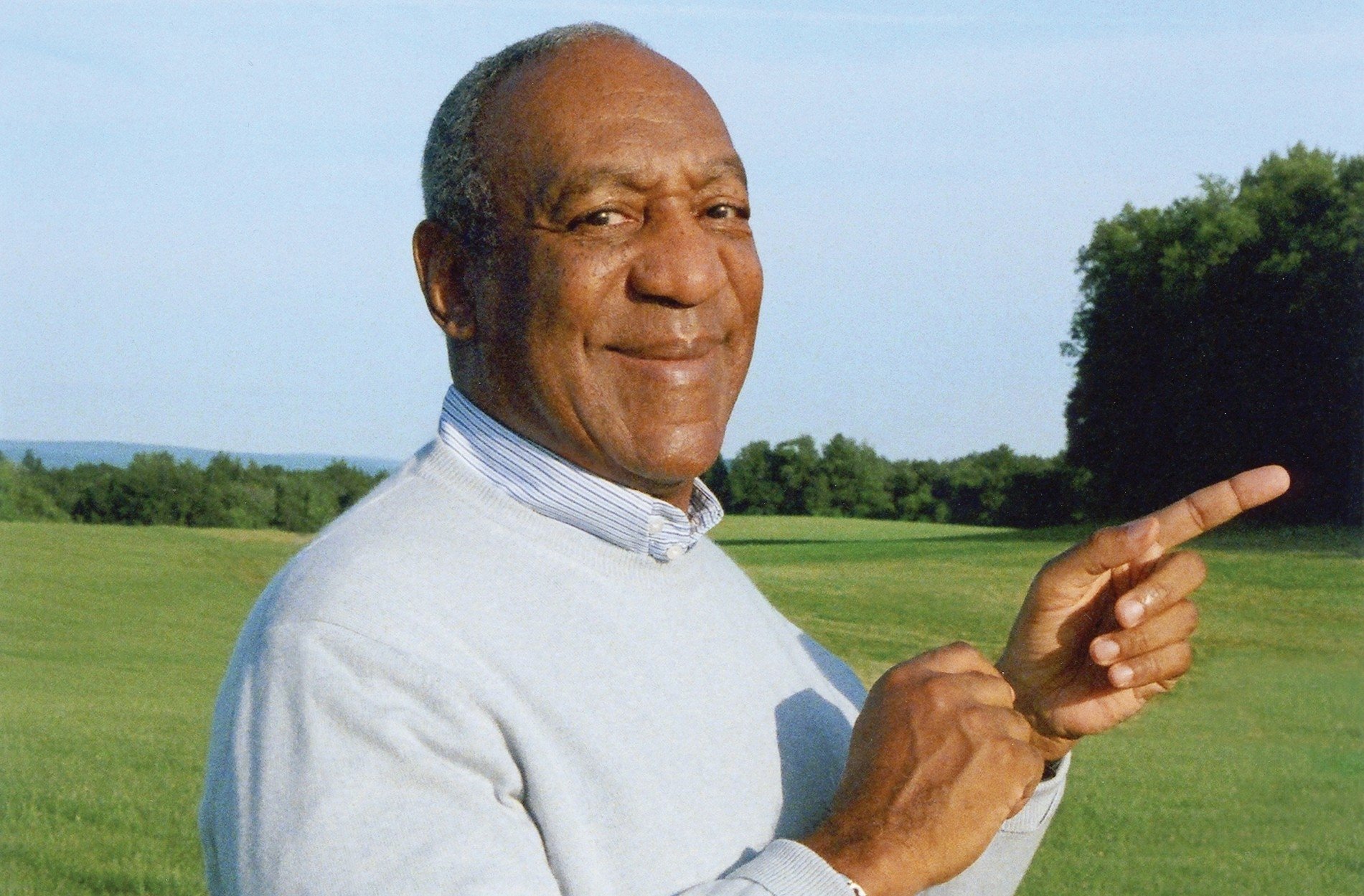 HD wallpaper of a smiling man in a white sweater pointing to his right, set against a green landscape.