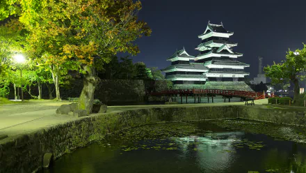 HD PC desktop background showing man-made Matsumoto Castle at night, brightly lit above a stone moat with reflections, framed by trees and a quiet park path.