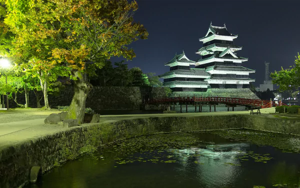 HD PC desktop background showing man-made Matsumoto Castle at night, brightly lit above a stone moat with reflections, framed by trees and a quiet park path.
