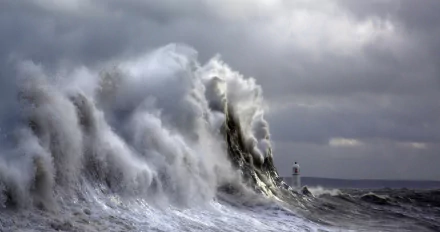 A powerful wave crashes against the shore near a lighthouse in Cardiff, Wales, under a dramatic stormy sky filled with clouds, capturing the wild beauty of nature.