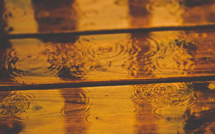 A close-up photograph of raindrops creating ripples on a wooden surface, capturing the tranquil beauty of rain in rich tones. This HD image serves as an engaging desktop wallpaper.