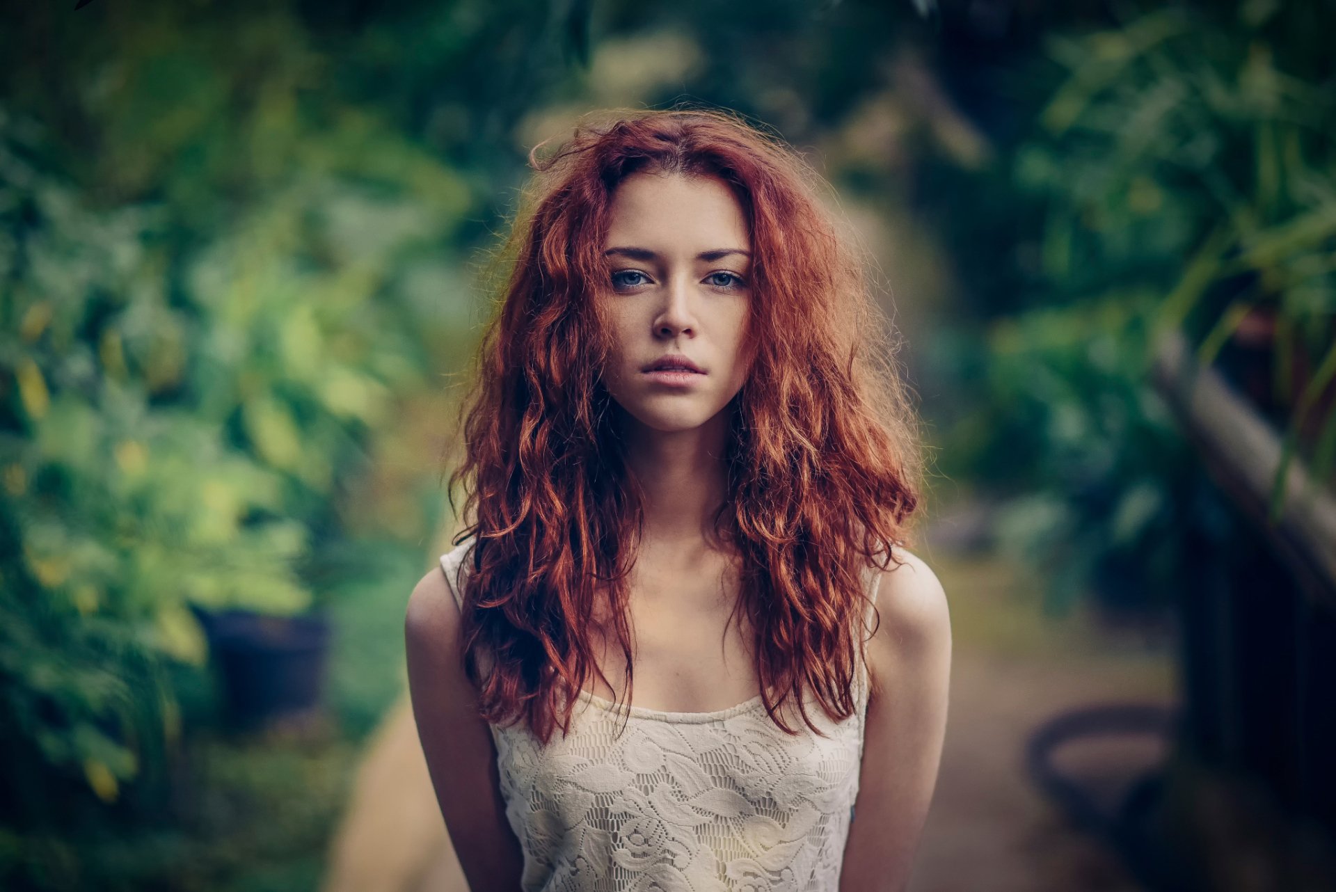 A redheaded woman with blue eyes stands confidently in an outdoor setting, her wavy hair framing her face. The background features a bokeh effect, adding depth to this HD desktop wallpaper.