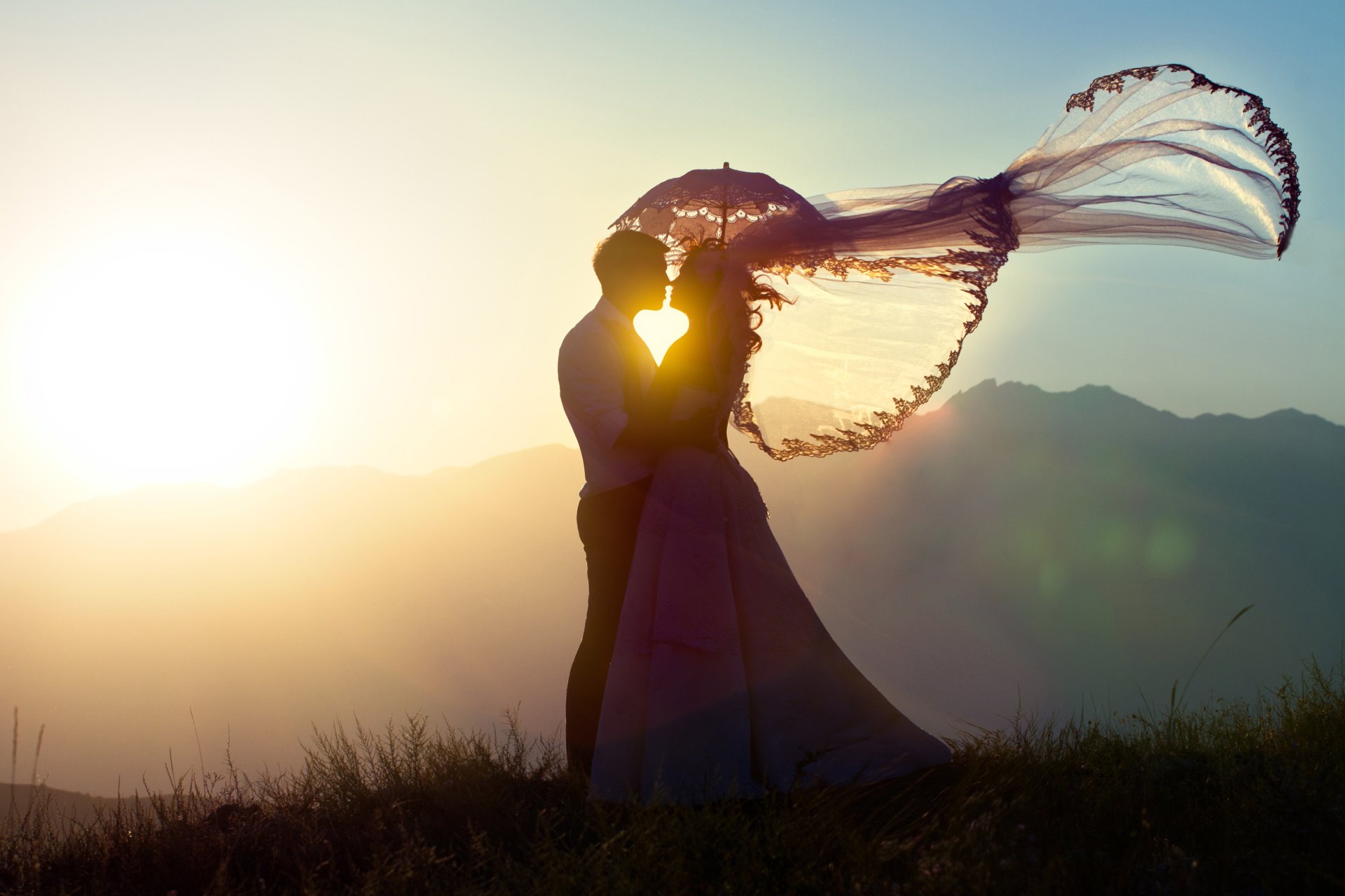 A couple embraces under a vibrant sunset, holding a flowing umbrella that captures a sense of love and warmth, set against a picturesque mountain backdrop. 4K Ultra HD wallpaper.