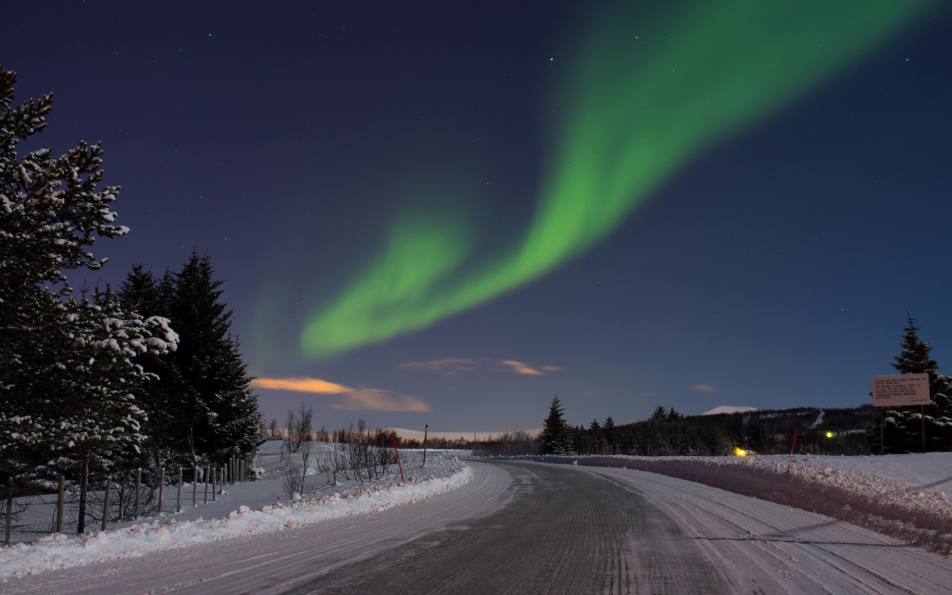 HD PC desktop wallpaper showing a snowy road winding through a forest at night beneath vibrant green aurora borealis lighting up the clear sky.