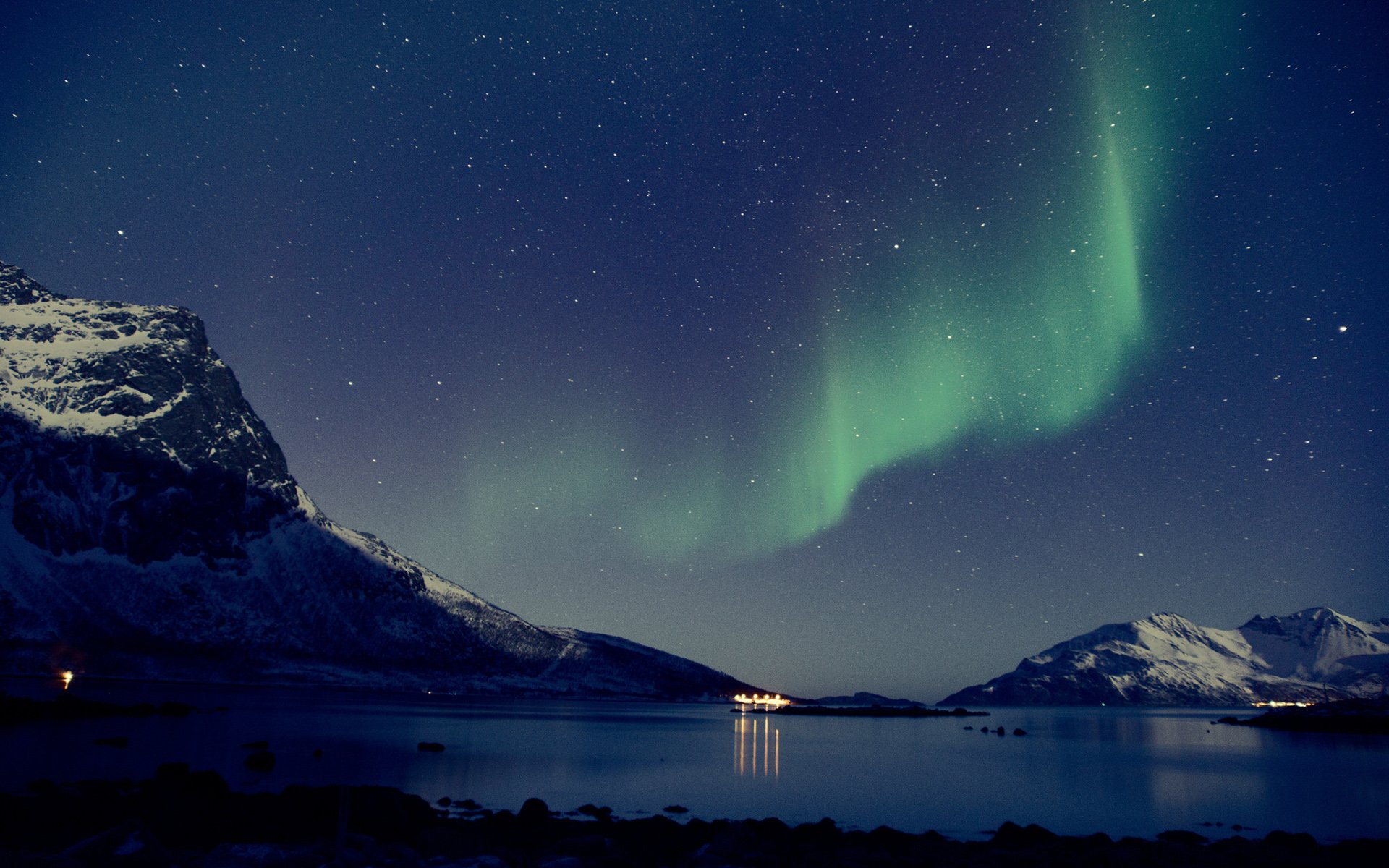 HD desktop wallpaper of a snowy mountain landscape at night, illuminated by the vibrant green glow of the aurora borealis over a calm lake.