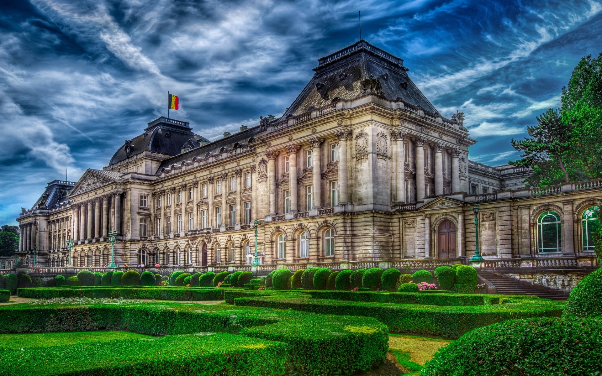 HD desktop wallpaper featuring the majestic Royal Palace of Brussels under a dramatic sky, showcasing its intricate architecture and manicured gardens.