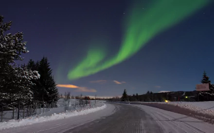 HD PC desktop wallpaper showing a snowy road winding through a forest at night beneath vibrant green aurora borealis lighting up the clear sky.