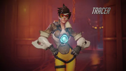 HD desktop wallpaper featuring Tracer from the video game Overwatch, standing confidently in her signature outfit with a glowing device on her chest. The background shows an industrial setting with warm lighting.