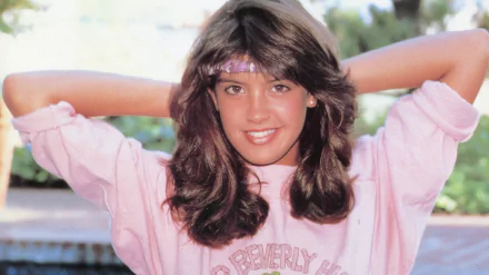 HD desktop wallpaper featuring celebrity Phoebe Cates smiling with hands behind her head, wearing a pink sweatshirt and a matching headband outdoors.