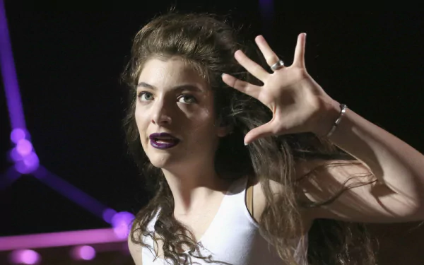 HD PC desktop wallpaper featuring singer Lorde with expressive hand gesture against a dark, purple-lit background, capturing her distinctive style and presence.