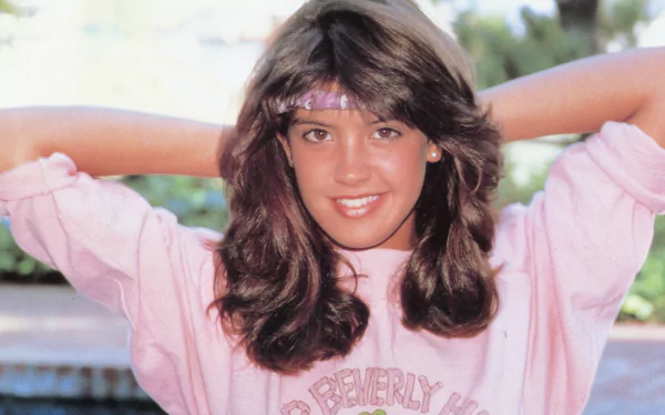HD desktop wallpaper featuring celebrity Phoebe Cates smiling with hands behind her head, wearing a pink sweatshirt and a matching headband outdoors.