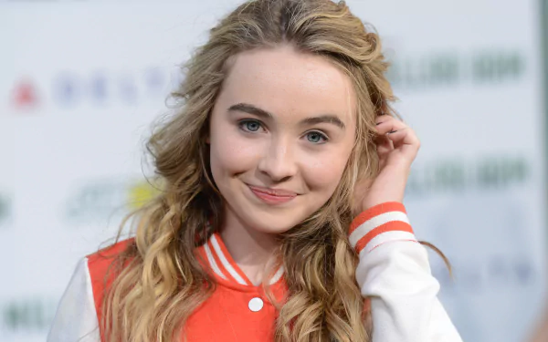 A blonde American actress with blue eyes smiles while wearing a red and white jacket. The background features a blurred event setting, creating a vibrant and inviting atmosphere.