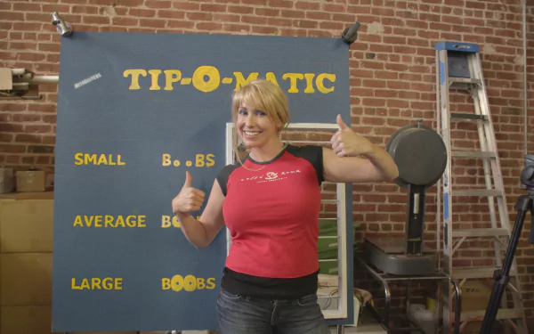 Mythbusters TV show 2K Quad HD PC desktop wallpaper: smiling woman gives thumbs-up in front of a TIP-O-MATIC board against a brick-walled workshop backdrop.
