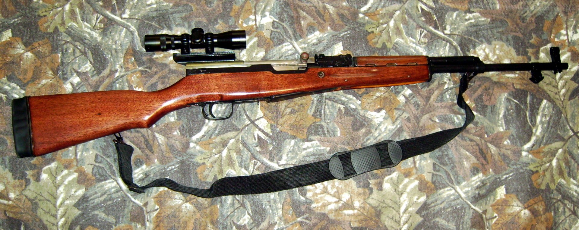HD PC desktop wallpaper featuring a man-made SKS rifle with wooden stock and attached scope, laid on a camouflage fabric background.