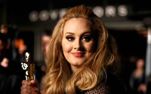 Adele, the renowned British singer, smiles while holding an award, captured in this HD wallpaper. The backdrop features blurred figures, enhancing her star presence.