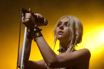 2K Quad HD PC desktop wallpaper of a blonde rock singer onstage holding a microphone, wrist cuffs and choker, bathed in warm yellow concert light.