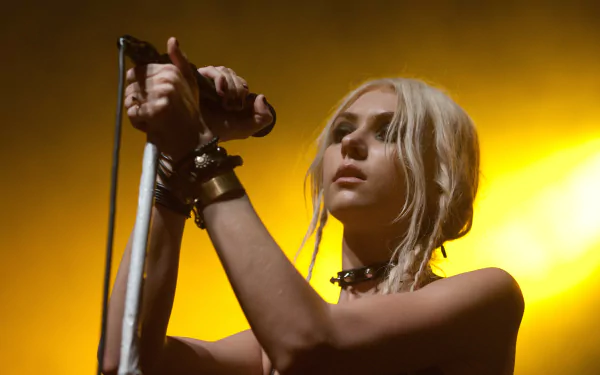 2K Quad HD PC desktop wallpaper of a blonde rock singer onstage holding a microphone, wrist cuffs and choker, bathed in warm yellow concert light.