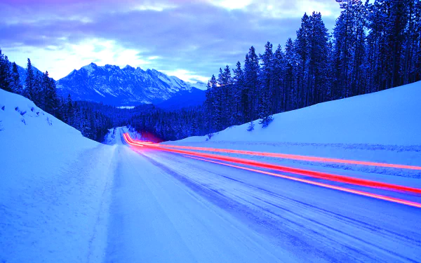 A snowy mountain road at twilight with tall trees along the sides, blue cold winter light, and red streaks from passing vehicles in this HD desktop wallpaper.