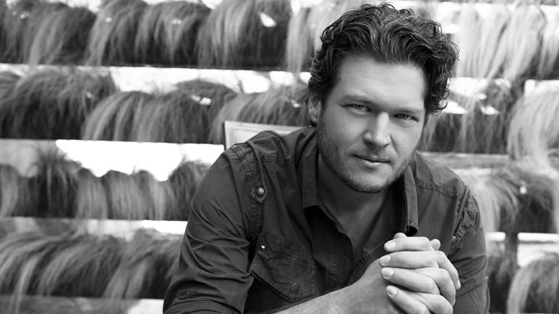 Black-and-white HD desktop wallpaper of a country music artist seated with hands clasped, wearing a casual shirt and wavy hair against a textured studio backdrop.