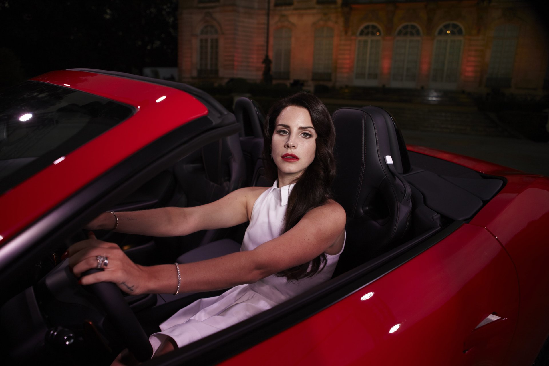 HD PC desktop wallpaper featuring Lana Del Rey seated in a red convertible, set against a nighttime urban backdrop, embodying a moody, cinematic music vibe.