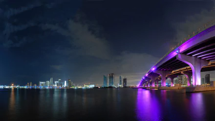 Night view of a man-made bridge in Miami illuminated with purple lights, reflecting on calm water, set against the city skyline in this HD PC desktop wallpaper.