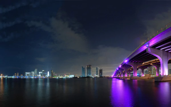 Night view of a man-made bridge in Miami illuminated with purple lights, reflecting on calm water, set against the city skyline in this HD PC desktop wallpaper.