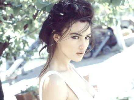 HD PC desktop wallpaper featuring celebrity Monica Bellucci posing outdoors with soft natural light filtering through leaves above.