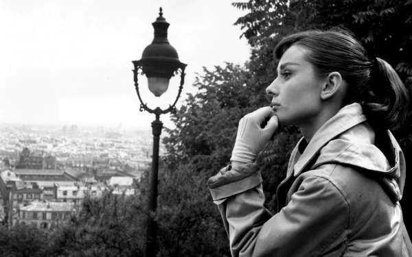 Black-and-white 2K Quad HD desktop wallpaper: classic celebrity portrait in profile, woman in a trench coat and gloves beside a vintage lamppost, gazing over a cityscape.