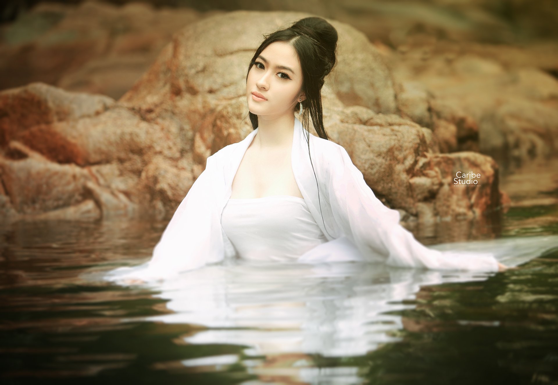 A Vietnamese woman in a white dress stands in calm water against a rocky background, captured in HD for a serene desktop wallpaper.