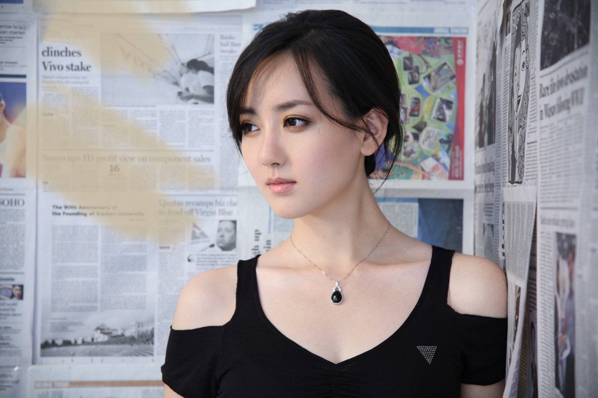 HD desktop wallpaper featuring an Asian woman, identified as Hú Yǐng Yí, standing against a backdrop of newspaper clippings. Hú Yǐng Yí is wearing a black top and a necklace, gazing thoughtfully into the distance.