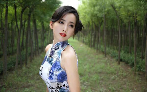 A poised Chinese woman named Hú Yǐng Yí stands gracefully in a lush green forest, wearing a floral dress, captured in a stunning HD wallpaper background.