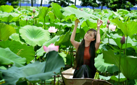 A Vietnamese woman sits in a boat on a pond surrounded by lush green lotus leaves and pink flowers, shielding herself with a large leaf. The serene scene captures the natural beauty of the environment.