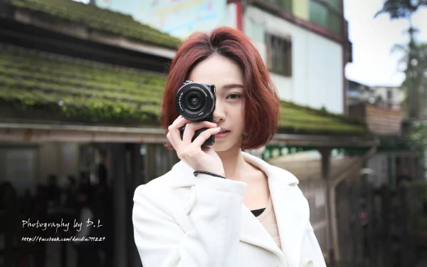 A Taiwanese woman named Lín Yǔ poses outdoors with a camera in this HD desktop wallpaper and background. She wears a white coat, and the setting features greenery and rustic buildings. Photography by B.L.