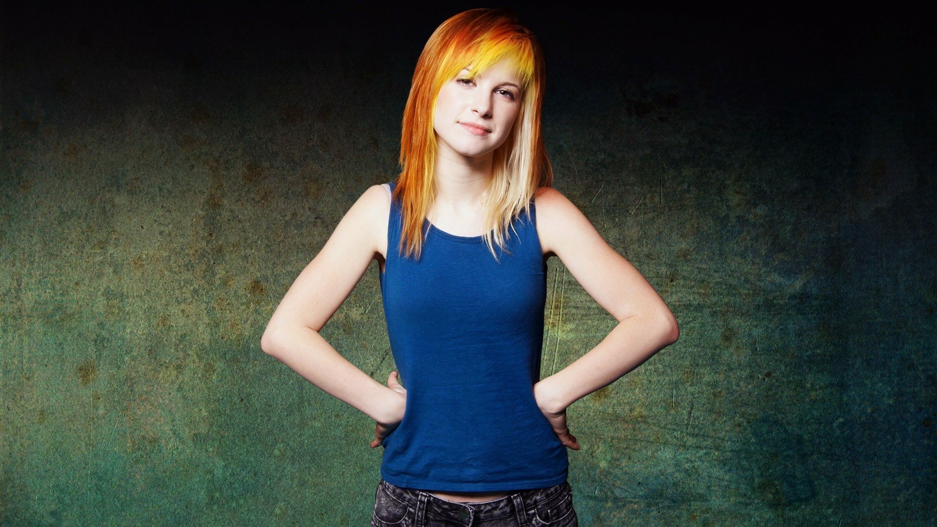 HD PC desktop wallpaper of a music artist with bright orange hair in a blue tank top, standing with hands on hips against a textured green backdrop.