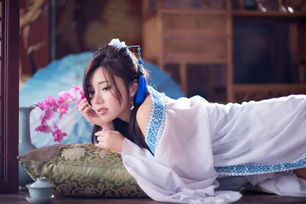 A woman in a flowing white dress rests on a cushion, surrounded by orchids and a vase, capturing an elegant Taiwanese aesthetic in this vibrant 4K Ultra HD wallpaper.
